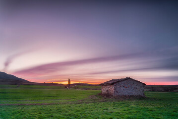 Naklejka premium Rustic stone hut in a green cereal field of Palencia at sunrise with a dramatic purple and orange sky over the rural landscape of Castilla y Leon