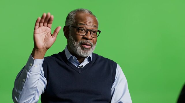 An elderly man waves his hand in a video recording studio with a green screen.