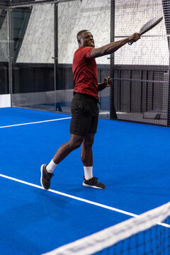 Male athlete swinging padel racket on blue court with glass walls, mesh netting and doodle panel