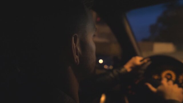 Close up of young guy holds hands on steering wheel and drives a modern automobile at evening. Male driver moves in his SUV car on empty road. Man rides in auto on highway at night time. Slow motion