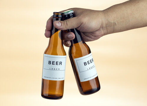 Close-Up of a Hand Gripping Two Handcrafted Lager Beer Bottles in Amber Glass With Clean Minimalist White Labels Showing 5.0% ABV Small Batch Brewing Details