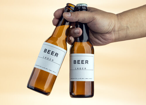 Close-Up of a Hand Gripping Two Amber Glass Handcrafted Lager Beer Bottles With Clean Minimalist White Labels Showing 5.0% ABV Small Batch Brewing Details