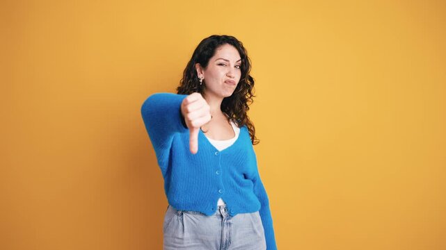Disappointed curly haired woman expressing dislike with a thumbs down gesture. Unhappy girl showing disapproval on a yellow background