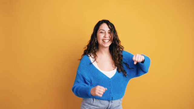 Joyful beautiful young woman with long curly hair dancing energetically against a yellow background. She is smiling and having fun