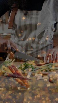 Vertical video: Cook placing knife dicing scallion, carrot on board, gold bokeh over hands cooking