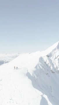 Vertical video: Pair appearing then traversing snowy ridge, pausing, continuing toward summit