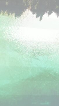 Vertical video: Turquoise lake shifting as sun glare growing triggers cam panning up showing forest