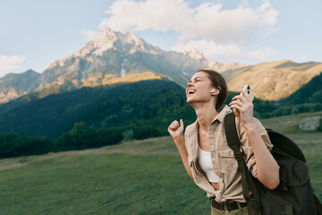 Naklejka premium A joyful woman hiking outdoors with a backpack smiles on a grassy hillside, with towering mountains in the background, capturing a moment of wanderlust.