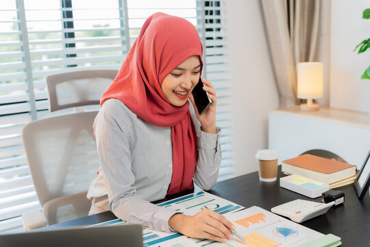 A Muslim businesswoman is working with financial graphs and a computer, remotely from a modern office.