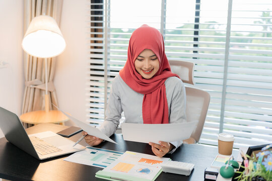 A Muslim businesswoman is working with financial graphs and a computer, remotely from a modern office.