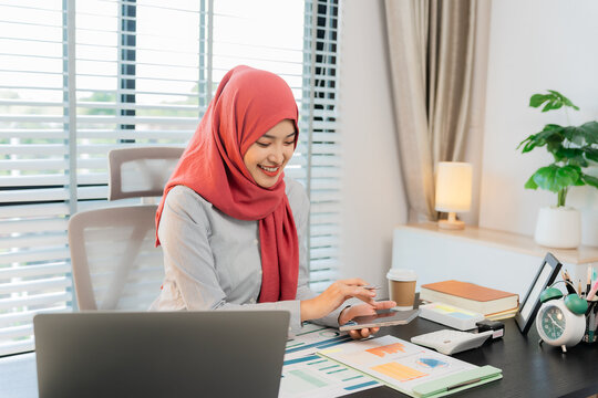 A Muslim businesswoman is working with financial graphs and a computer, remotely from a modern office.