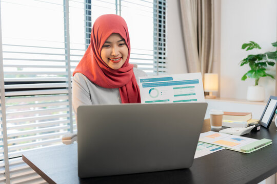 A Muslim businesswoman is working with financial graphs and a computer, remotely from a modern office.