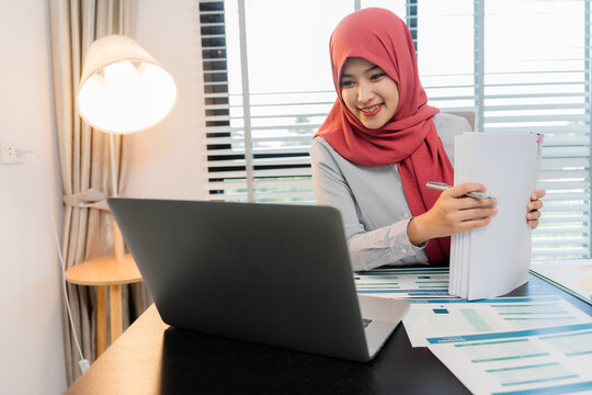 A Muslim businesswoman is working with financial graphs and a computer, remotely from a modern office.