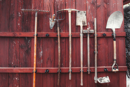 Vintage garden tools hanging on a weathered red wooden wall, with rake, shovel, hoe, pickaxe and fork arranged in rustic farm storage