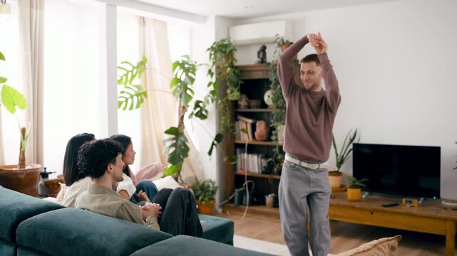 Young man acting and gesturing while playing pantomime game with friends. His cheerful companions sitting on the sofa and guessing the word