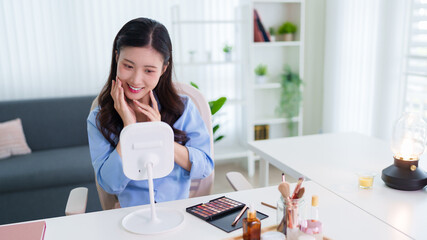 young Asian woman applying makeup using lighted mirror table at home living room modern beauty...