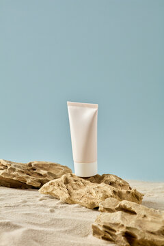 Minimalist cosmetic mockup: white tube on natural stones and sand, blue background. Perfect for organic skincare branding and beauty product presentation.
