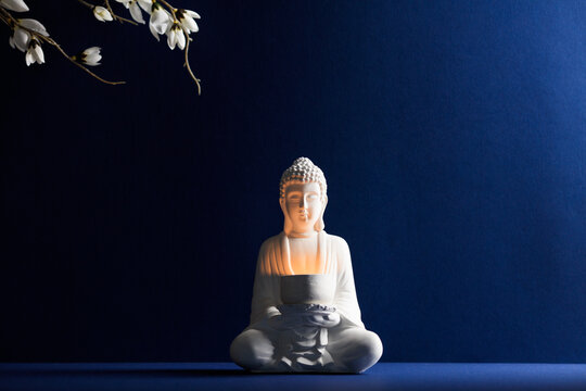 Central white Buddha statue in meditation pose with a glowing bowl, framed by delicate white blossoms on a navy blue background. Spiritual enlightenment concept.