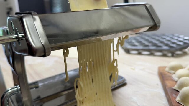 Fresh pasta dough pressed through stainless steel pasta machine, thin noodle strands falling onto wooden counter, home kitchen prep scene with rustic cooking mood