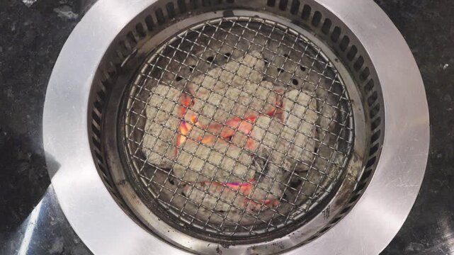 Top view of hot charcoal glowing under a wire mesh grid in a circular BBQ grill. Stainless steel restaurant equipment for Japanese yakiniku or Korean tabletop barbecue style dining.