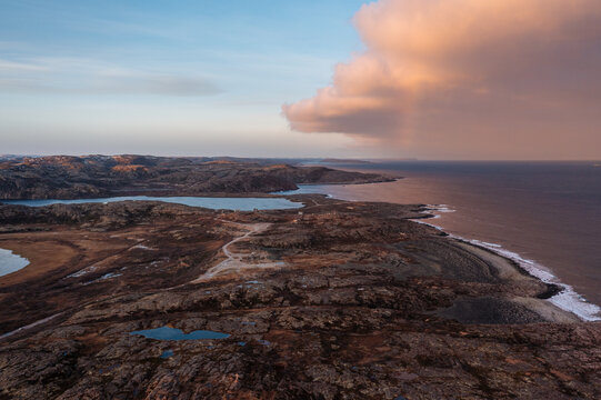 Aerial landscape of the Barents Sea coast featuring rugged tundra, small lakes, and a massive pink cloud at sunset. Arctic Ocean.