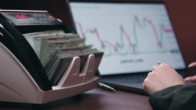Professional analyzing financial charts while counting machine handles stack of cash nearby. Concept business performance and capital tracking.
