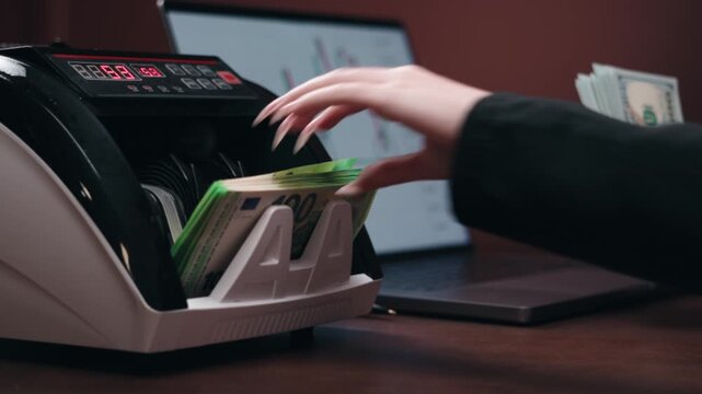 Female financial analyst operates a currency note counting machine while reviewing data on a laptop, stacks of cash visible on a wooden desk in a modern office setting