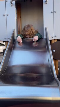 Curly hair child playing on slide at playground. Excited kid preparing for descent on metal chute. Active toddler enjoying outdoor game in public park setting.