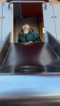 Curly hair child playing on slide at playground. Excited kid preparing for descent on metal chute. Active toddler enjoying outdoor game in public park setting.