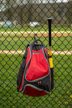 A red baseball equipment bag with a bat hangs from the chain link fence at a youth baseball field, the green grass of the outfield visible.