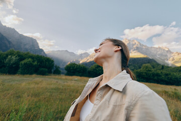 Naklejka premium A woman in a white shirt stands in a wide field, mountains in the distance, looking up at the sky. A lifestyle moment conveying nature, calm, and outdoor freedom.