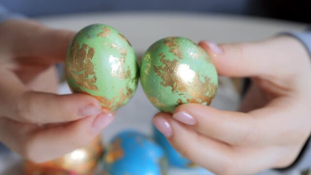 Close-up of hands holding two green and gold decorated Easter eggs. Background shows blurred colorful eggs. Focus remains on the detailed egg surface texture