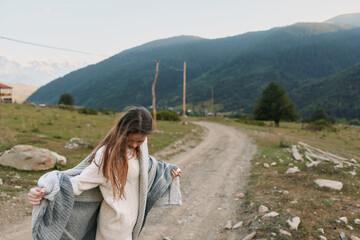Fototapeta premium Young woman with long hair wraps a gray shawl around her shoulders while walking on a dirt road in a mountainous rural area during a calm day.
