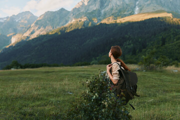 Naklejka premium A woman with a backpack stands in a grassy meadow, looking toward distant mountains, embodying outdoor life, hiking, and solitude in nature.