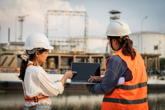 Two engineers collaborate near a construction site. One points at a laptop screen while the other types. White hard hats and safety vests signal professional readiness