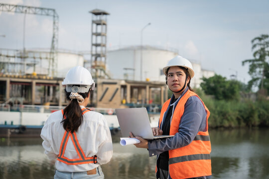 Two engineers stand by a waterway near industrial structures. One holds a laptop, the other carries rolled plans. Both wear helmets and high-visibility vests