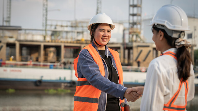 Two workers shake hands at a construction site. They wear hard hats and reflective vests. One smiles warmly, showing team spirit. The background features buildings and water