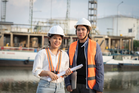 Two engineers stand by a waterfront industrial site. They wear hard hats and safety vests, smiling at the camera. One holds blueprints, the other carries a tablet