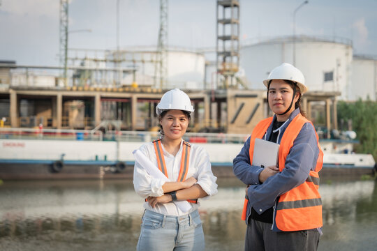 Two women in hard hats stand confidently beside a waterway. They wear reflective vests, facing the camera with calm assurance. Industrial tanks and structures loom behind them, reflecting in the water
