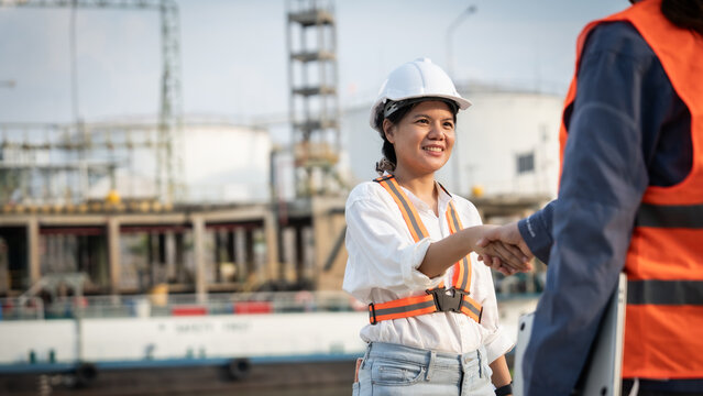 Smiling woman shakes hands with colleague at construction site. Both wear safety gear, showing teamwork and trust. Background shows steel frames and industrial structures