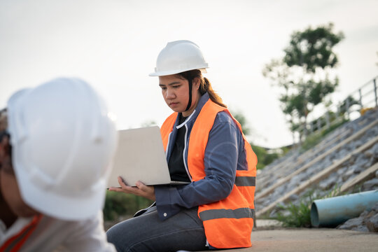 Engineer sits with laptop, reviewing plans. Wearing hard hat and high-visibility vest. Focused, attentive, engaged with construction task. Outdoor site shows pipes and concrete structures