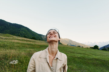Naklejka premium Cheerful woman outdoors in a beige jacket, smiling upward in a sunlit meadow with distant mountains, soft breeze, and vibrant greenery surrounding.