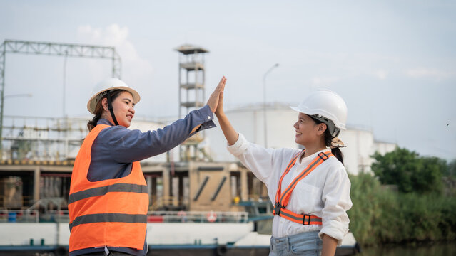 Two workers high-five, celebrating teamwork. Hard hats and vests show safety compliance. Industrial backdrop suggests construction or maintenance. Their smiles hint at success and mutual respect