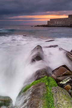 Sunset at Punta Galea Getxo with dramatic orange clouds over a rocky coast and long exposure silk effect on the ocean waves near the port wall