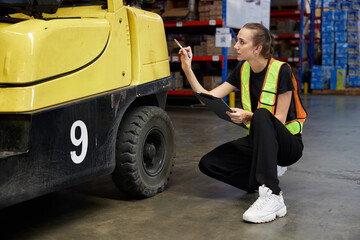 Worker inspecting a forklift and writing on a clipboard in a warehouse © offsuperphoto