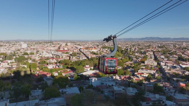 Aerial drone footage of a vibrant red cable car gondola gliding over Durango Mexico cityscape with sprawling urban buildings, green trees, and distant mountain range under clear blue sunny skies.