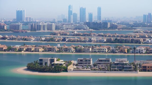 Dubai aerial view showcasing Palm Jumeirah island with luxury waterfront villas and Mediterranean style homes. Turquoise waters surround the fronds while city skyscrapers rise in the hazy distance.