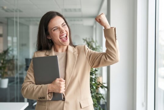 Happy businesswoman holding a laptop, raising fist, and cheering, showing achievement and victory