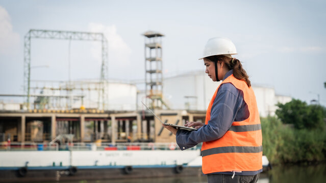 Woman in hard hat and vest uses tablet outdoors. Industrial facility looms behind her in background. She appears focused, reviewing data on her device