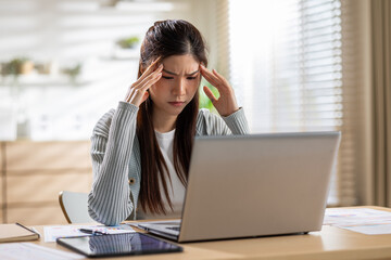 Frustrated Asian woman experiences severe headache and stress while working on laptop computer at home office desk feeling overwhelmed and exhausted from excessive screen time and heavy workload